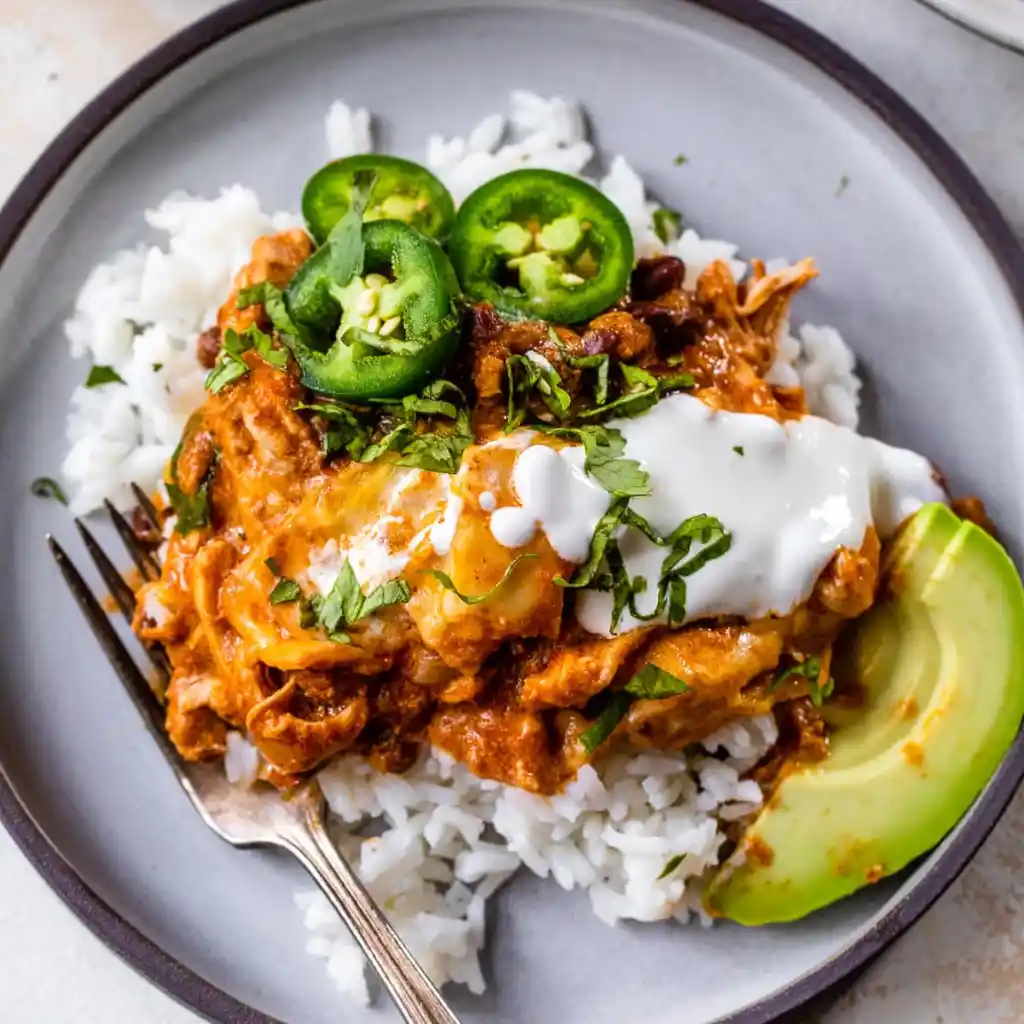 Slow Cooker Chicken Enchiladas 12 Plate of slow cooker chicken enchiladas served over rice with avocado, sour cream, and jalapeños.