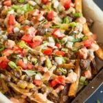 omemade cheeseburger fries on a baking tray topped with ground beef, melted cheese, diced tomatoes, onions, lettuce, and creamy sauce.