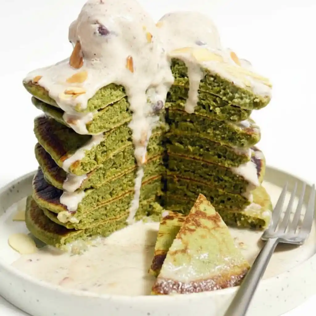 A tall stack Japanese Fluffy Matcha Pancakes Recipe topped with melting ice cream and almond slices, served on a white plate with a fork.