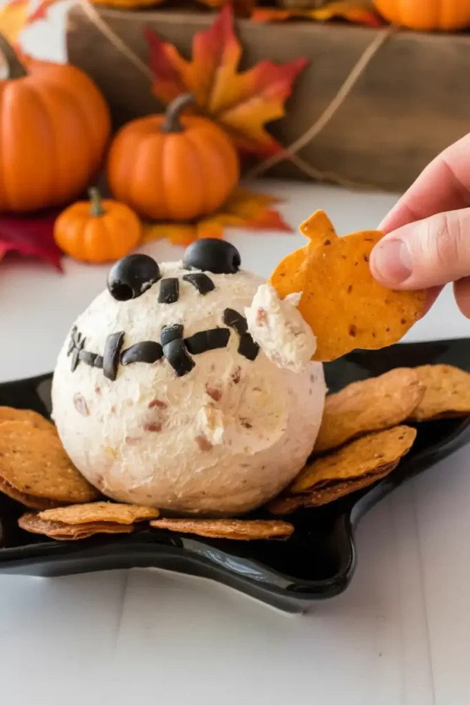 Skellington Cheese Ball Appetizer Recipe 4 A hand dips a pumpkin-shaped cracker into a Skellington cheese ball appetizer. The appetizer is presented on a black plate with crackers and a festive fall background featuring pumpkins and autumn leaves, highlighting a perfect Skellington Cheese Ball Appetizer Recipe.