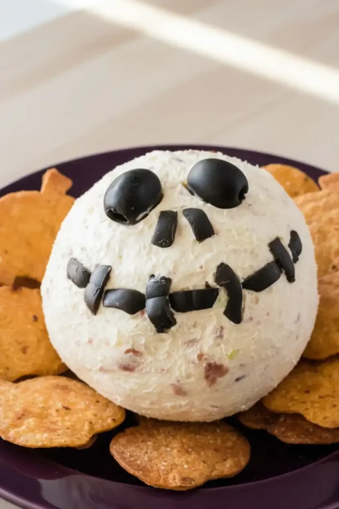 Skellington Cheese Ball Appetizer Recipe 3 A close-up of a Skellington cheese ball appetizer, with a face made of black olives, served on a purple plate and surrounded by pumpkin-shaped crackers. This image showcases a fun and easy Skellington Cheese Ball Appetizer Recipe, perfect for Halloween.